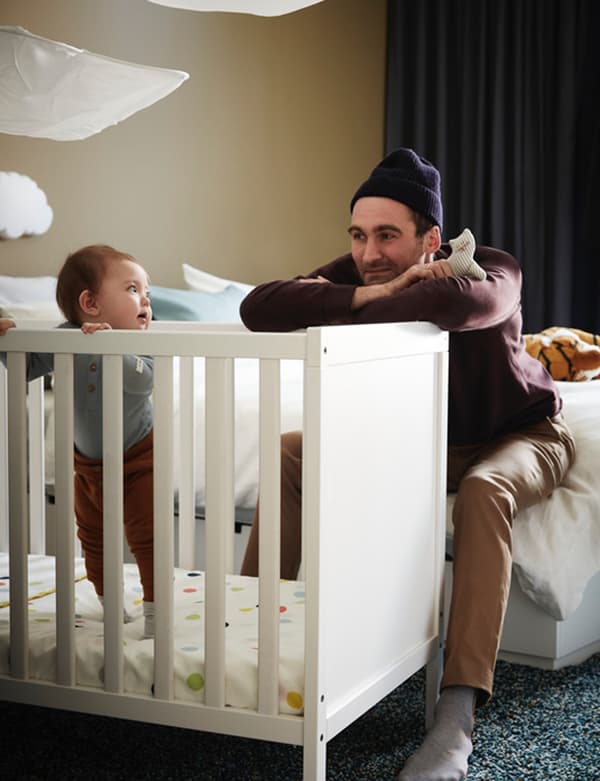 Father looking at son who is standing in an IKEA SUNDVIK crib with spotted bed sheet in a bedroom.
