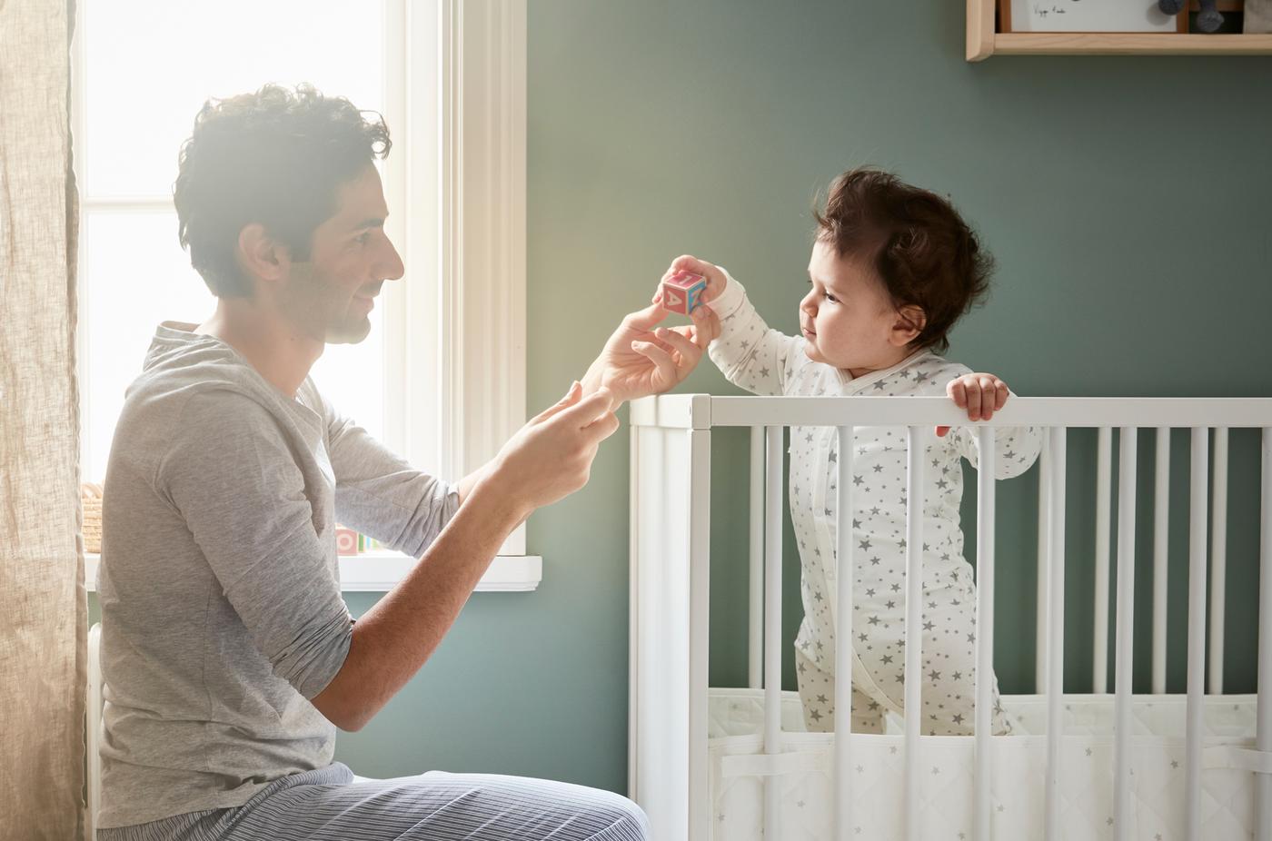 Father playing a game with his baby and a small wooden toy as the baby stands in a white cot holding on to the balustrade.