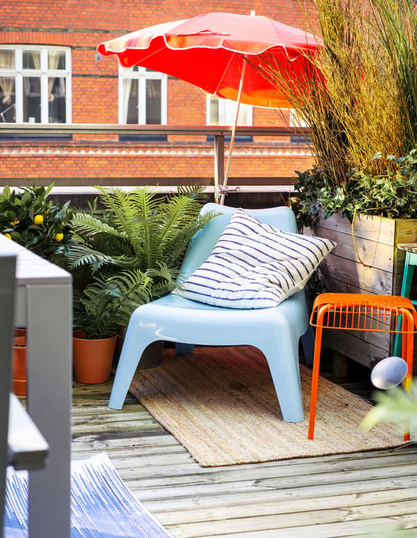 Fauteuil bleu dans le coin d’un balcon, entouré de plantes vertes, avec grand parasol.