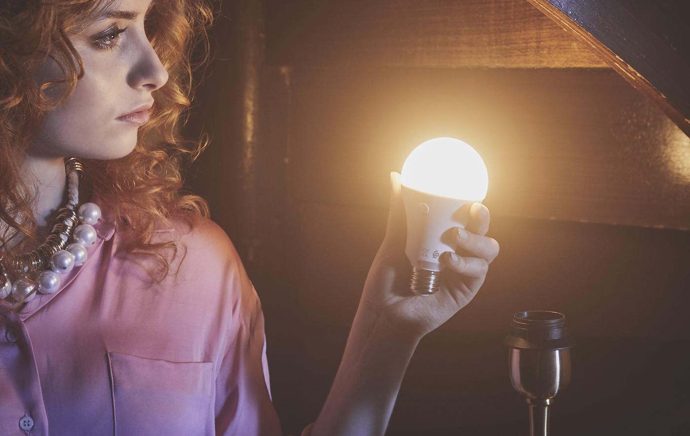 Femme à la chevelure rousse tenant en main une ampoule allumée, dans une pièce revêtue de lambris.