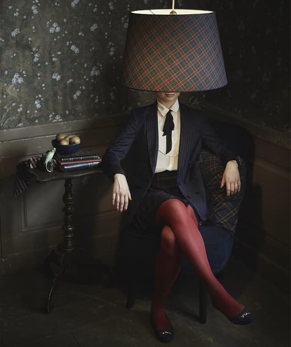 Femme assise dans un fauteuil, dans une pièce sombre, avec un abat-jour à motifs de carreaux à la place de la tête.