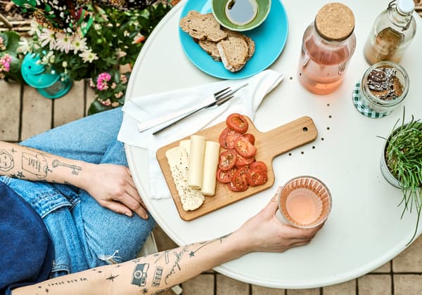 Femme aux bras tatoués assise à une table ronde couleur crème. Elle tient un verre, à côté d’une planche de fromage et de tomates.