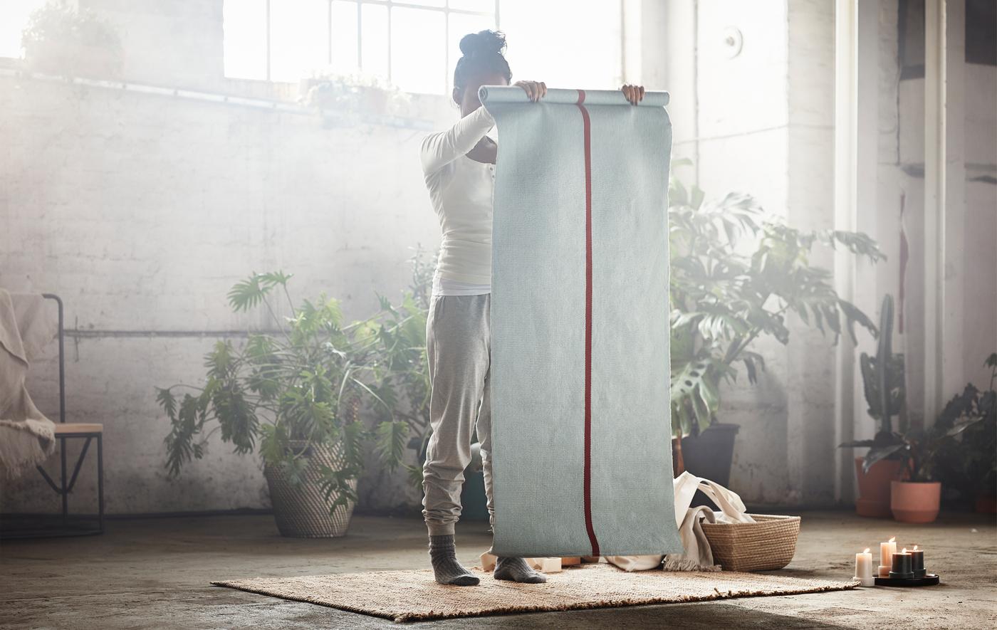 Femme debout dans un entrepôt et entourée de plantes vertes, tenant en main un tapis de yoga.