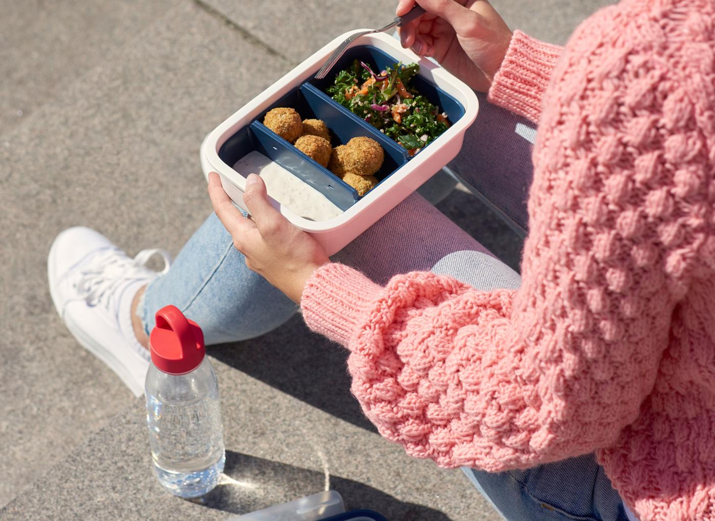 Femme en train de manger dans un récipient alimentaire en plastique IKEA, avec une bouteille en plastique transparent remplie d'eau derrière elle.