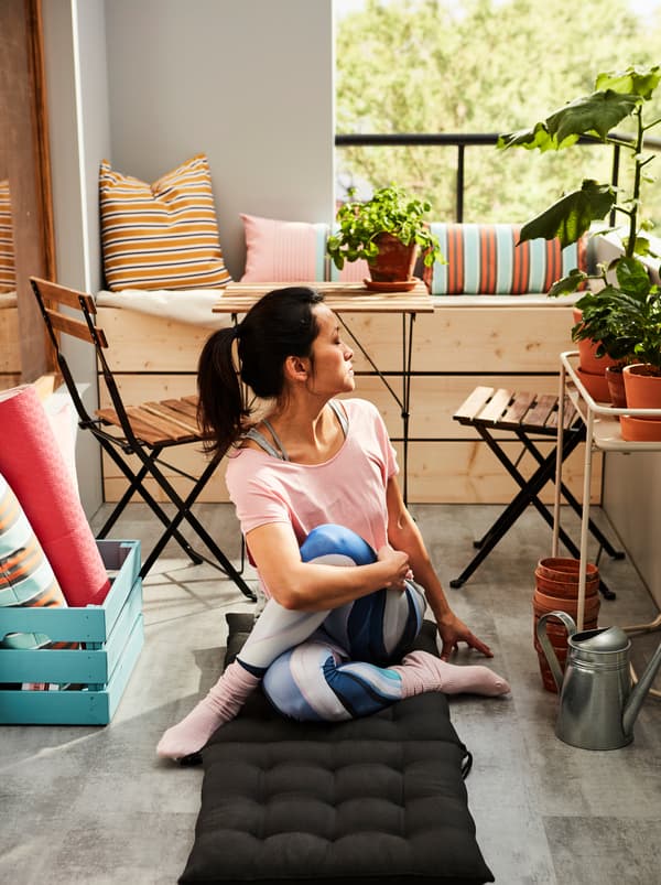 Femme prenant une pose de yoga, installée sur un coussin d’assise/de dossier HÅLLÖ posé sur le sol d’un balcon ensoleillé.