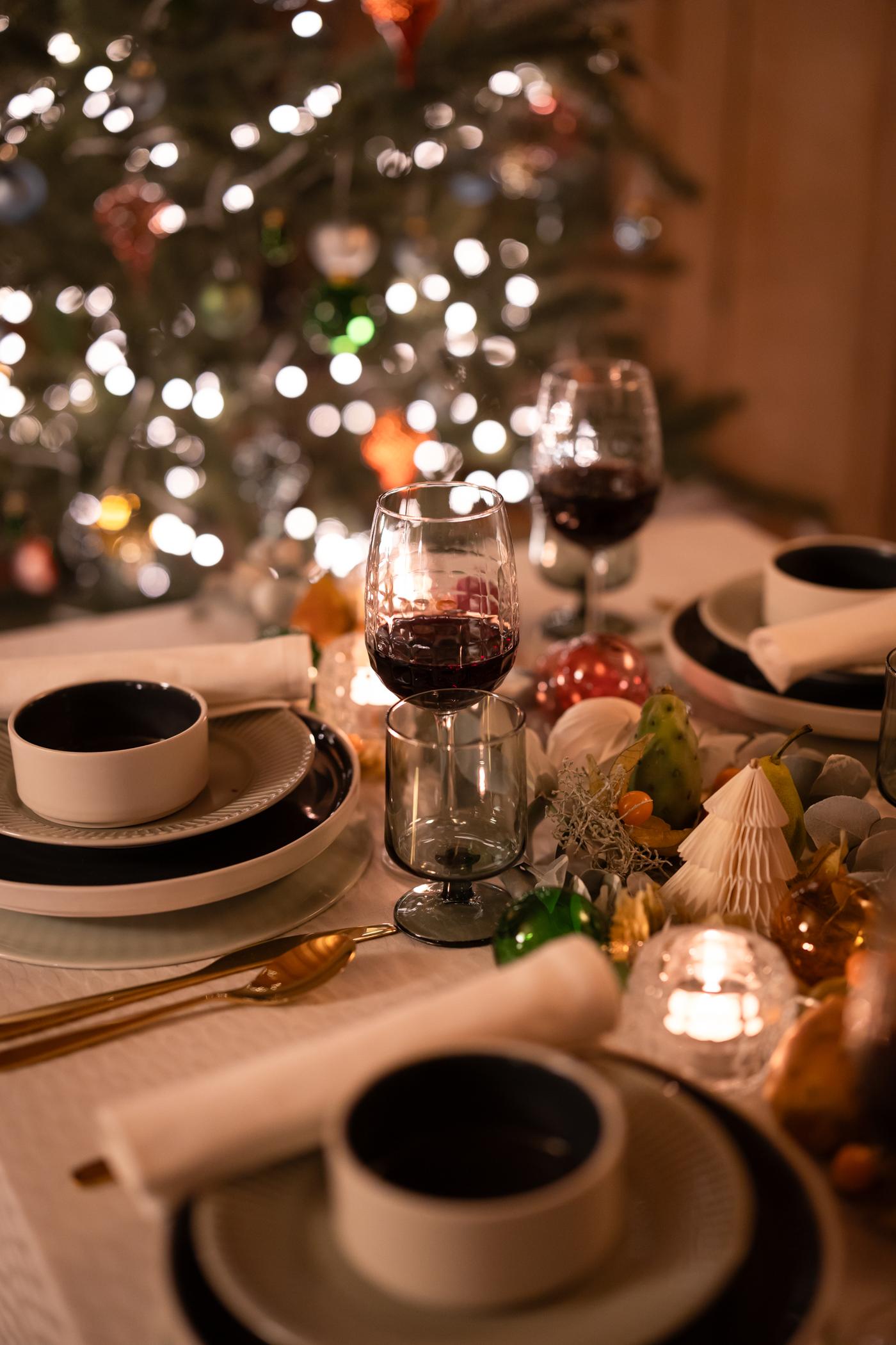 Festive dining table set with elegant dishware, wine glasses, gold cutlery, and holiday decorations; a lit Christmas tree in the background adds warmth.