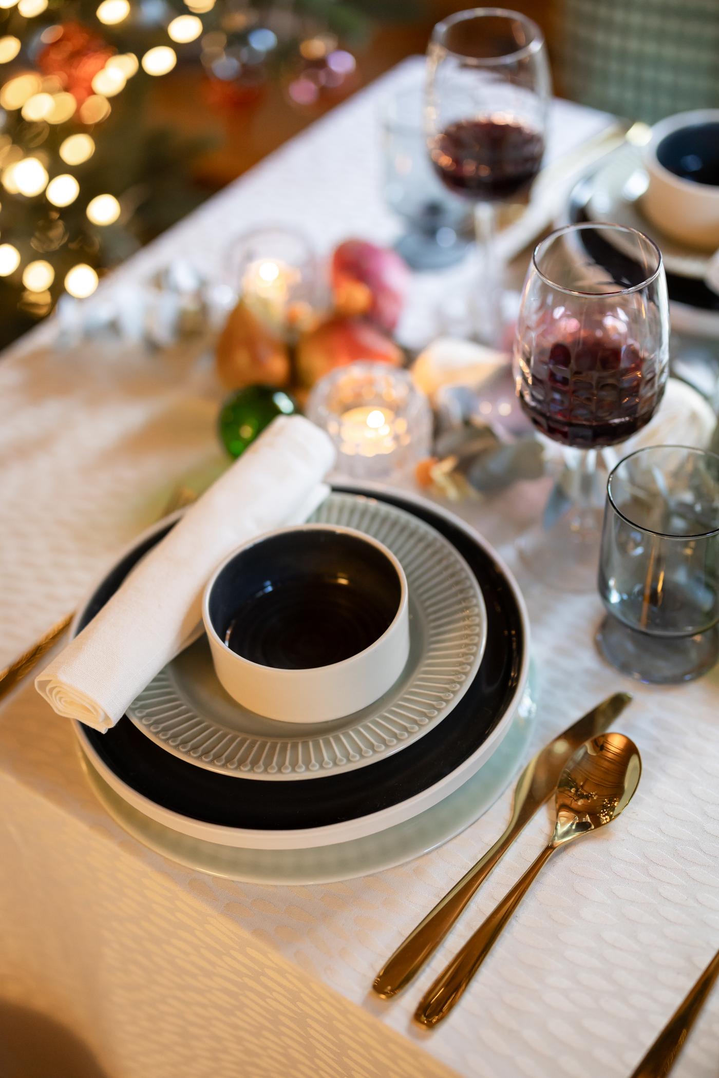 Festive dinner table with elegant place setting: stacked plates, a bowl, folded napkin, gold utensils, wine glasses, and soft candlelight. Cozy holiday ambiance.