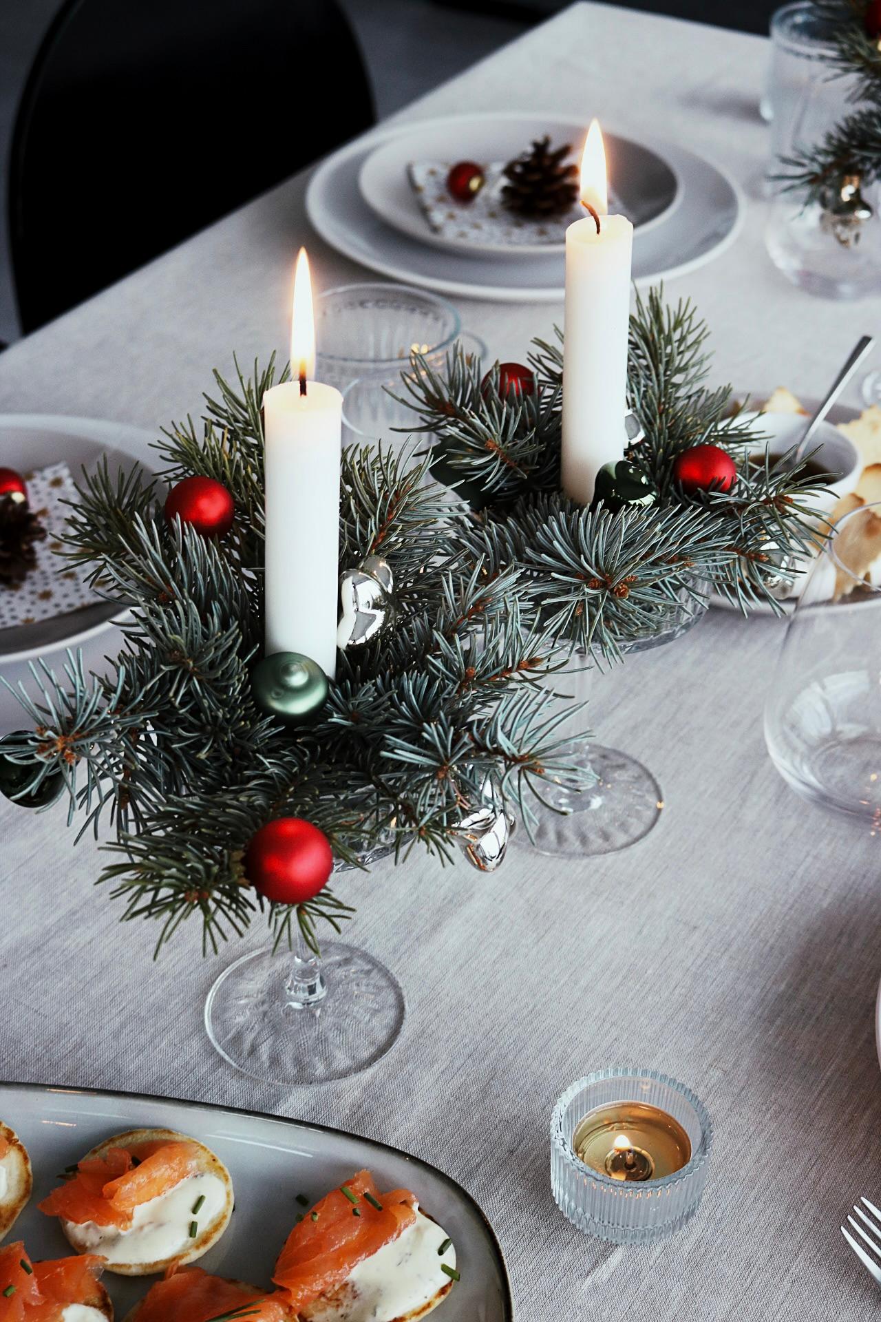 Festive table setting with lit candles in greenery wreaths, red and green ornaments, and plates of appetizers. Cozy holiday ambiance.