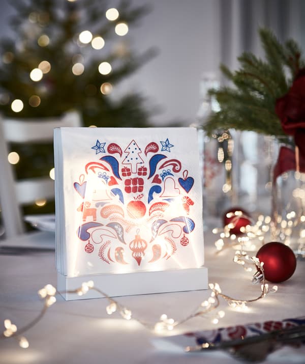 Festively patterned paper napkins standing upright in a lit stand, thus adding to the holiday-set table it’s placed on.