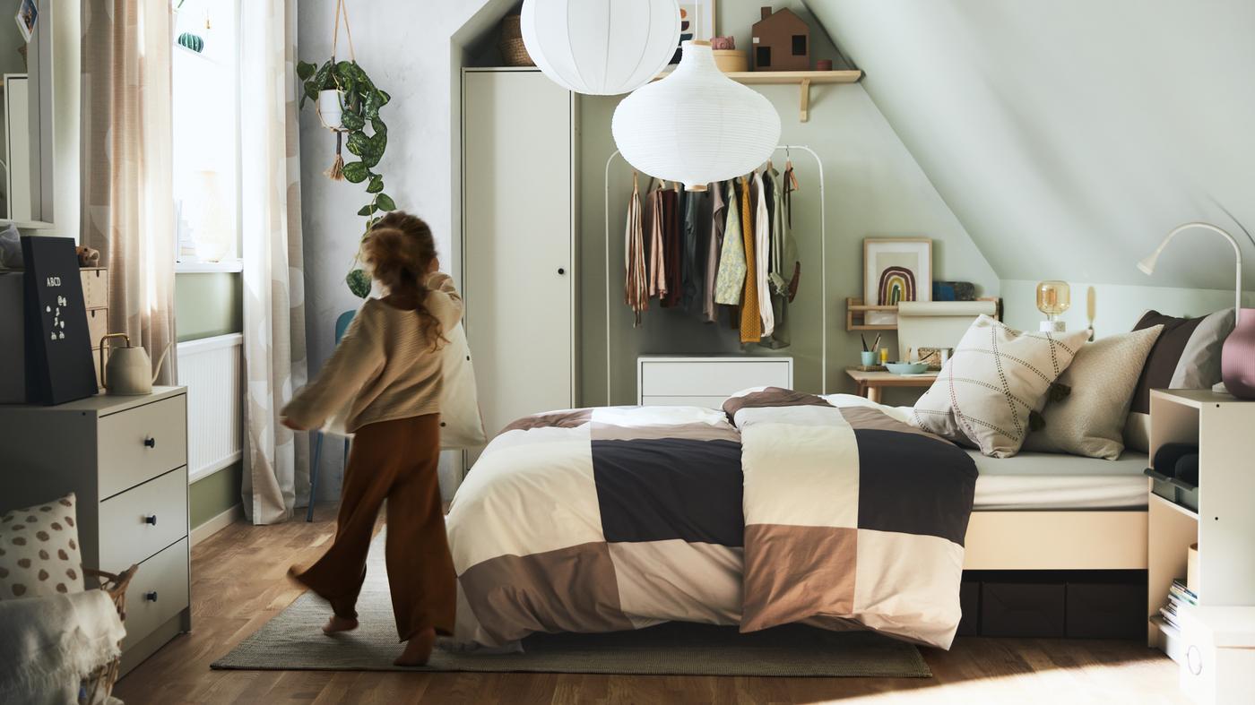 Fillette entrant dans une chambre meublée d’un lit, avec du linge de lit à carreaux brun/gris, de rangements et de suspensions blanches.
