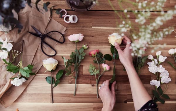Fleurs artificielles et fleurs coupées variées, alignées sur une table en bois, à proximité de ciseaux et de rouleaux de ruban adhésif.
