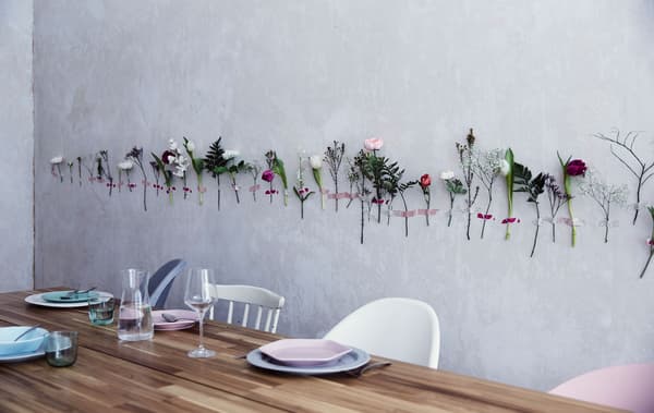 Fleurs sur tige fixées au mur à l’aide de ruban adhésif. En avant-plan, table en bois, assiettes et couverts.