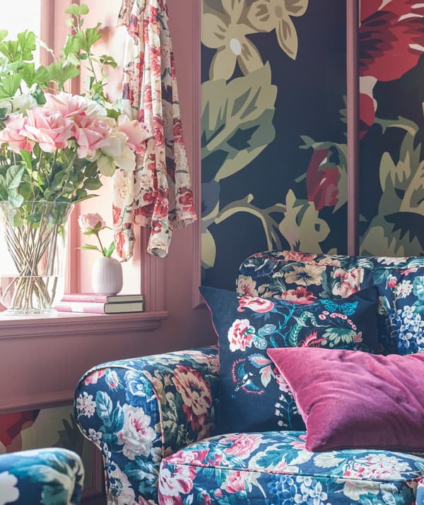 Floral patterned sofa with deep pink cushion in a room decorated with bold floral wallpaper and a pink window frame.