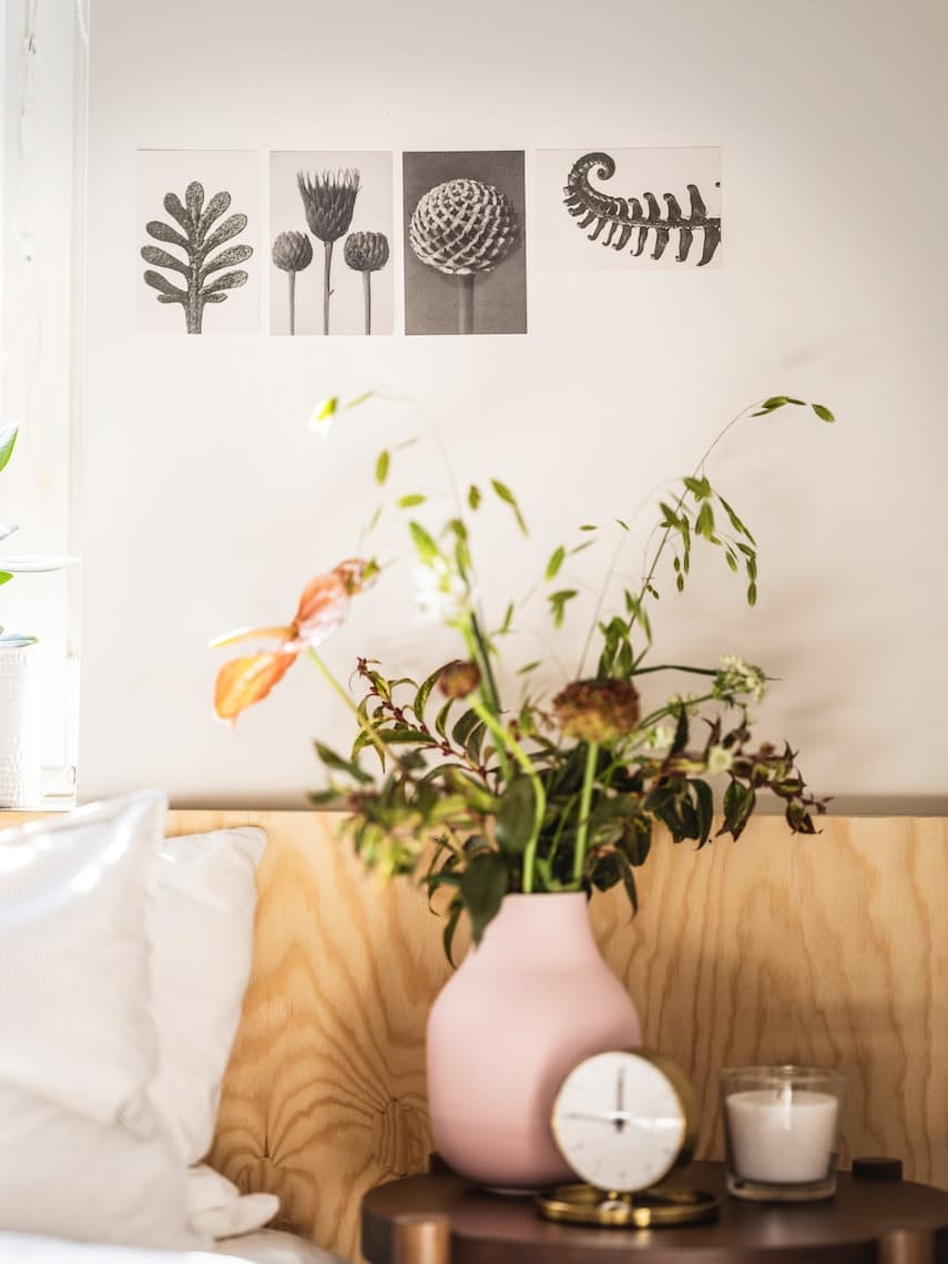 Florero de color rosado con planta en su interior junto a un reloj y una vela.