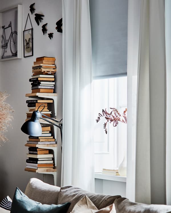 Fotografía de una ventana con cortinas blancas, y una estantería llena de libros con una lámpara al lado.