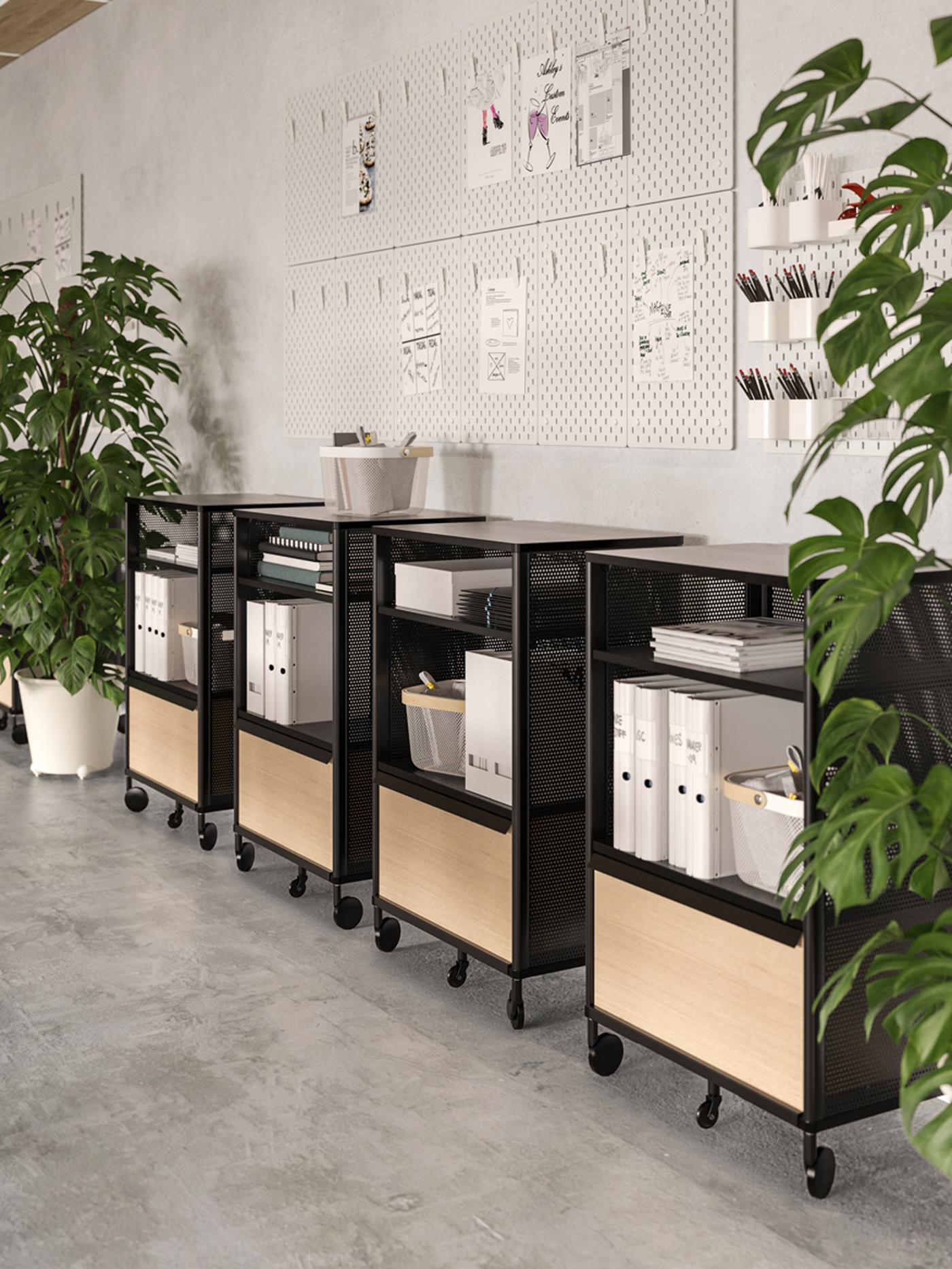 Four black BEKANT storage units with smart lock and castors between two potted plants, against a wall with pinned documents.