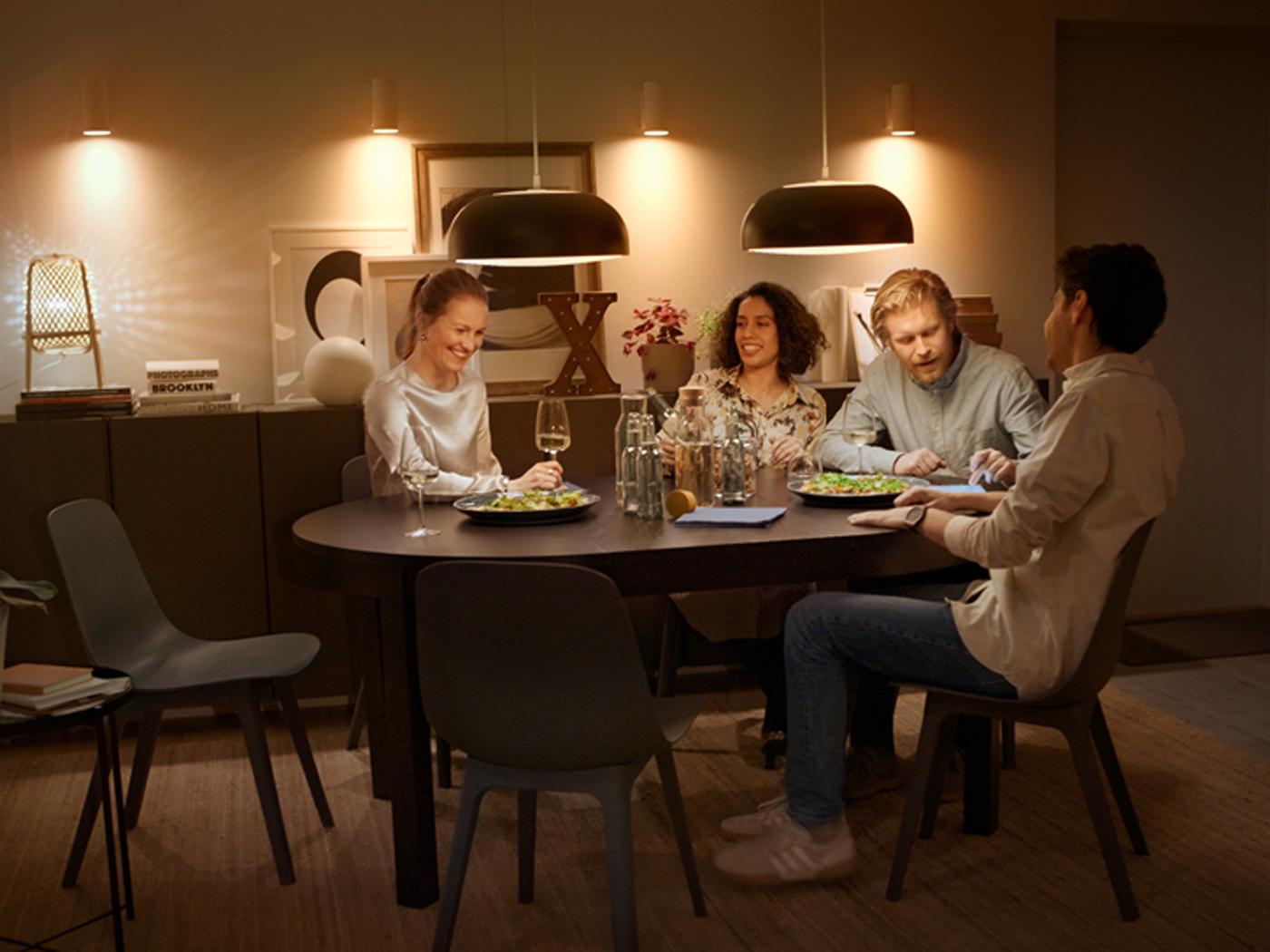 Four friends eat at a BJURSTA extendable table under pendant lamps controlled by a system for smart light and more at home.