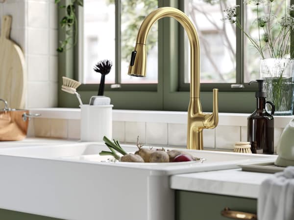 Fregadero de cocina moderno con grifo dorado, gabinetes verdes, cepillos de limpieza, dispensador de jabón líquido y verduras frescas en la barra.