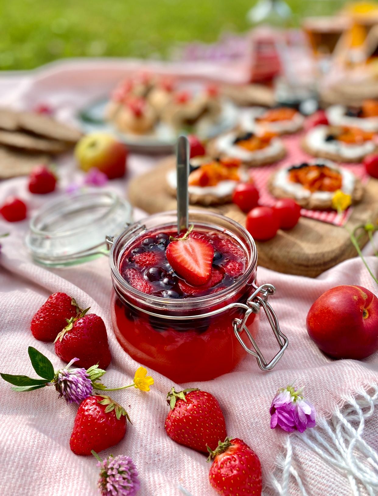 Frische Erdbeermarmelade in einem Glasgefäss mit einem Löffel, umgeben von ganzen Erdbeeren und einer Picknickkulisse im Hintergrund.
