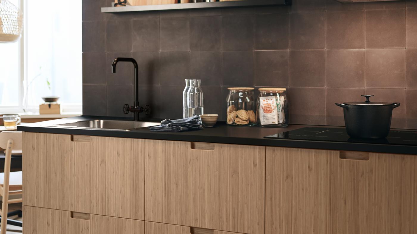 FRÖJERED kitchen fronts in bamboo, with diverse glass jars on the work surface and a cooking pot on the stove.