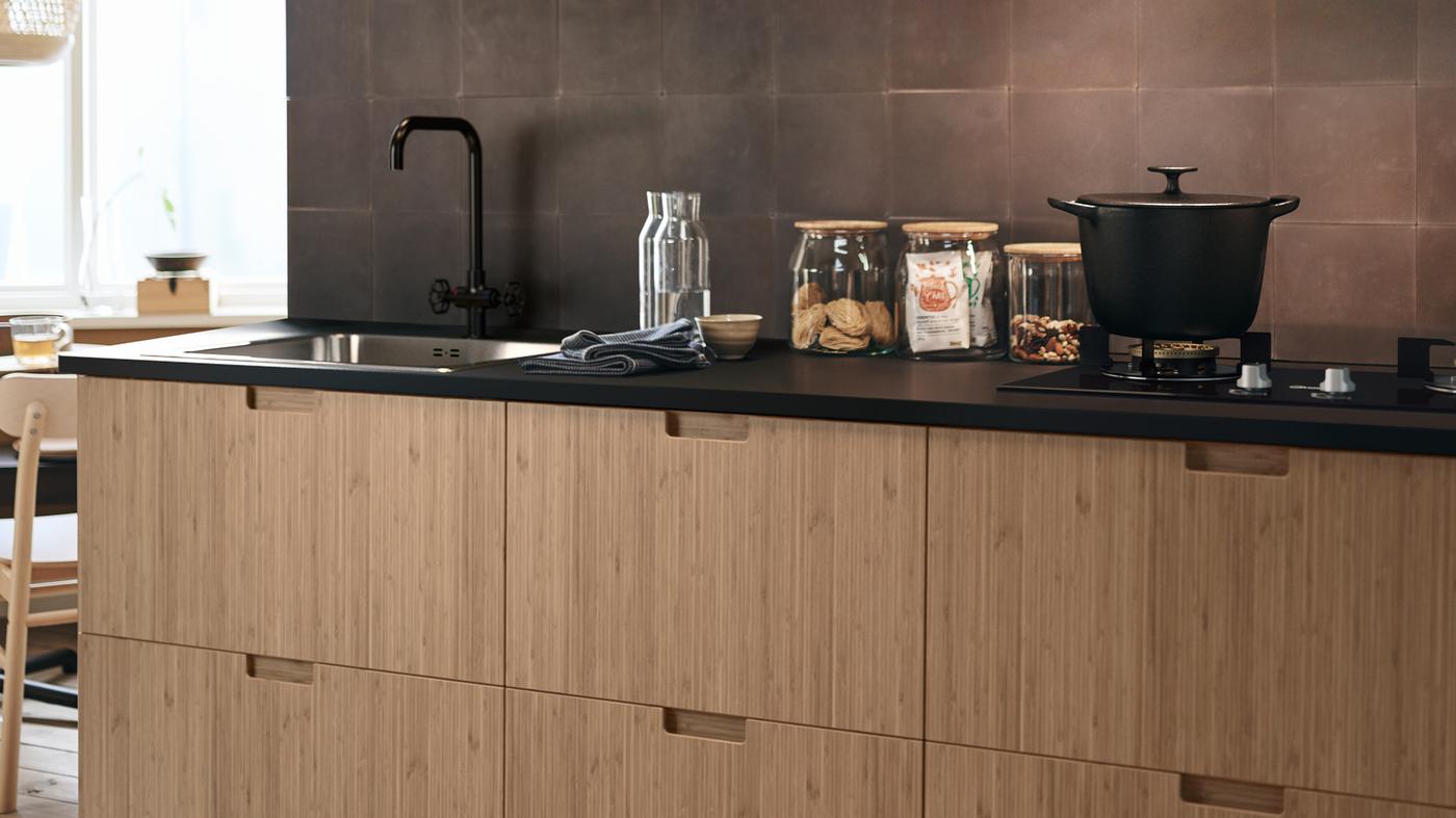 FRÖJERED kitchen fronts in bamboo, with diverse glass jars on the work surface and a cooking pot on the hob.