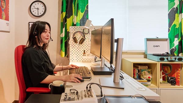 女性がデスクの赤オフィスチェアに座っている。PC作業している。 