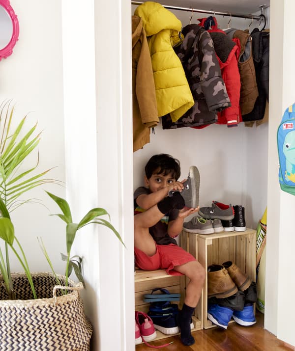 Garçonnet en train de mettre ses chaussures, assis sur un cageot en bois, en dessous de manteaux sur cintre dans une armoire.