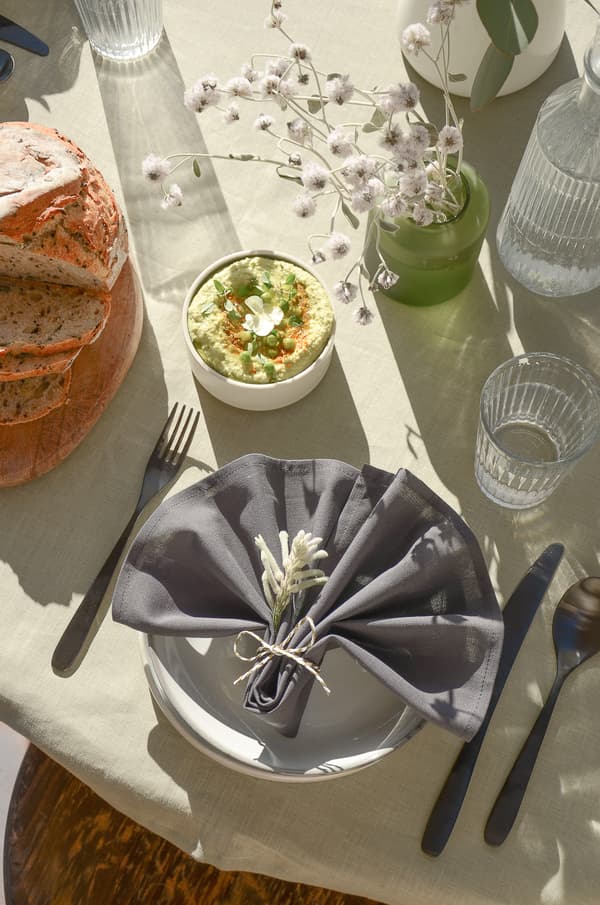Gedeckter Tisch mit Brot, Dip, grauer Serviette mit Blumen, Gläsern und Trockenblumen auf heller Tischdecke.