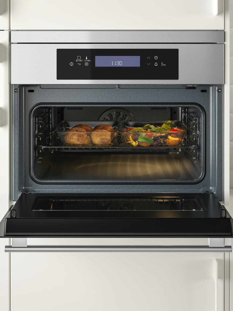 Geöffneter Backofen mit digitalem Bedienfeld oben; im beleuchteten Innenraum befindet sich ein Blech mit mehreren Lebensmitteln, die gerade gegart werden.