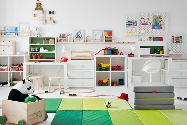غرفة أطفال بها ألعاب على الأرض، ومكاتب على الجنب، ورسومات على ألواح تعليق ورفوف للصور FLISAT على الجدران من أعلى.