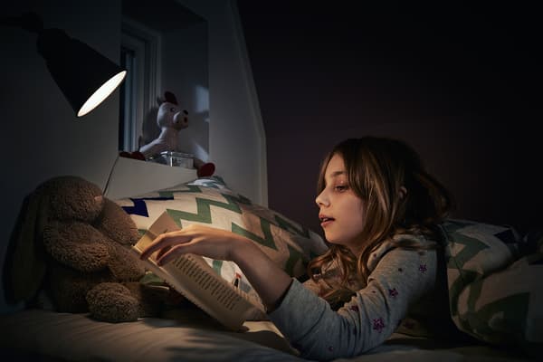 Girl wearing pyjamas reading a book in bed using a small lamp.