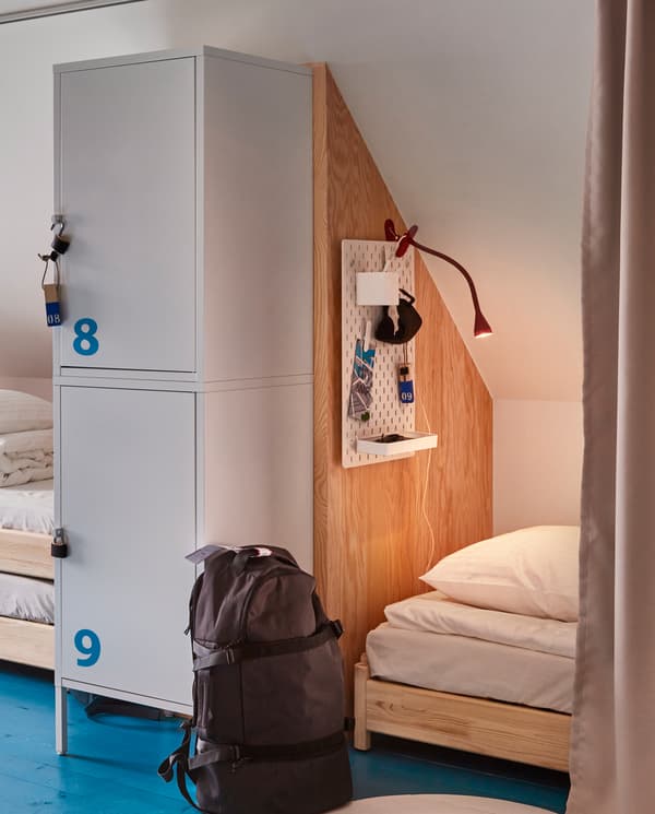 Glimpses of two hostel beds, separated by a wall and storage lockers, in the front there’s a black backpack.