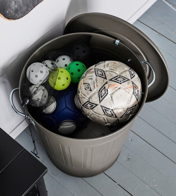 Grand bac gris avec couvercle ouvert montrant un ballon de football et de petites balles en plastique dans un filet noir.