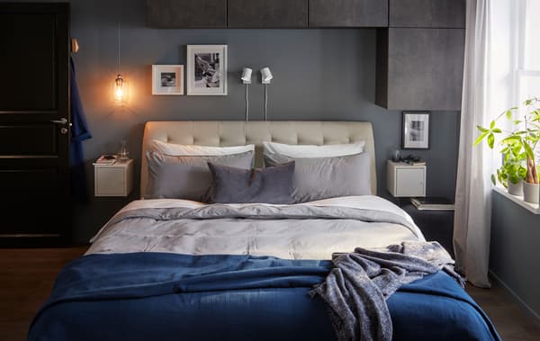 Grand lit double dans une chambre à coucher aux couleurs gris-bleu. Eléments muraux au-dessus et à côté du lit, plantes sur l’appui de fenêtre.