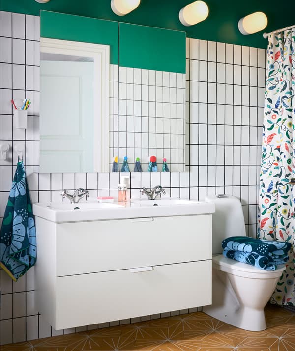 Grand miroir installé au-dessus d’un meuble-lavabo double dans une salle de bains aux murs vert et carreaux blancs.