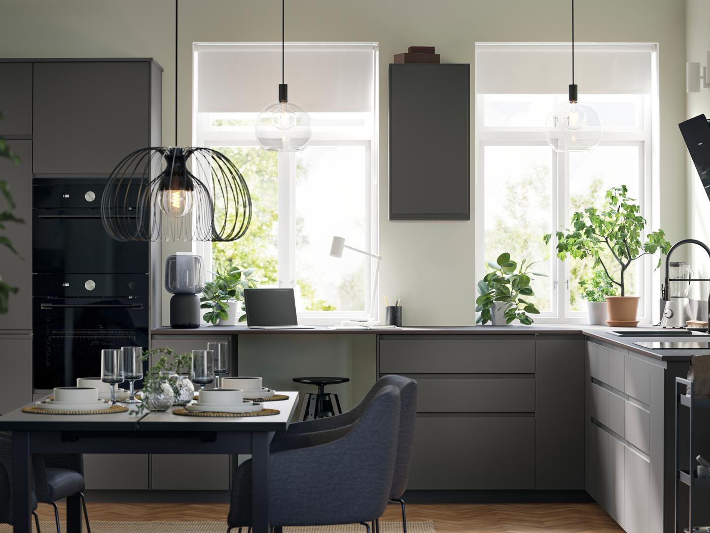 Grande cuisine avec façades VOXTORP gris foncé, électroménagers noirs, deux grandes fenêtres et un espace salle à manger.