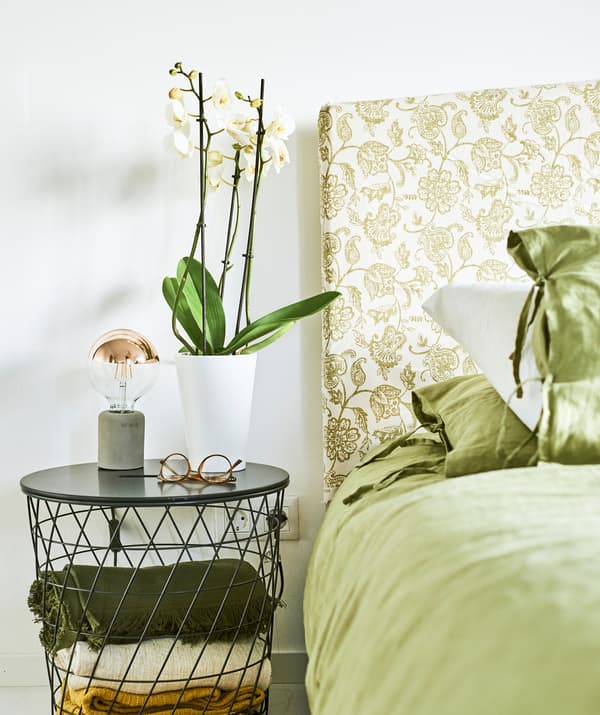 Green bed linen on a bed with a headboard made of green paisley fabric, specs and an orchid sit on the wire bedside table.