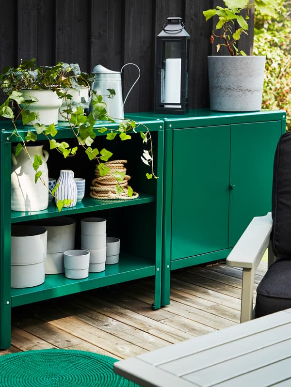 Green storage units outside, holding various pots, jugs and plants in plant pots, a lantern and some table mats.