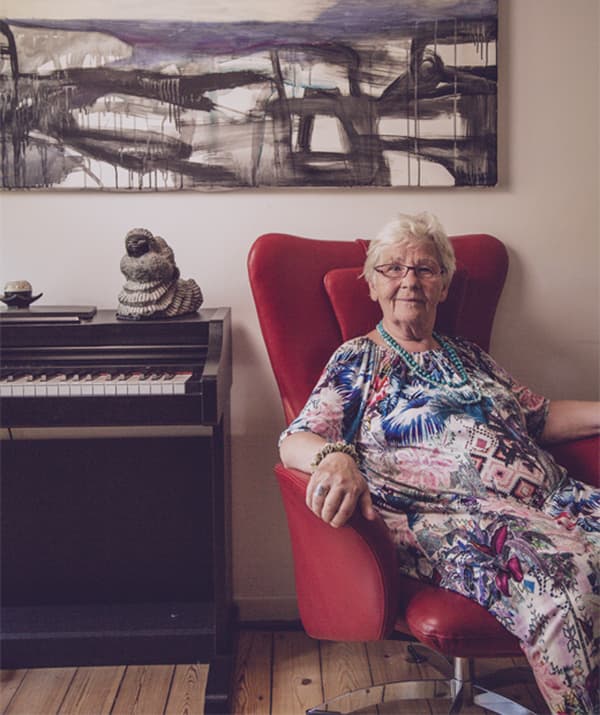 Greta sits in a red winged chair next to a piano with artwork on the wall behind her.