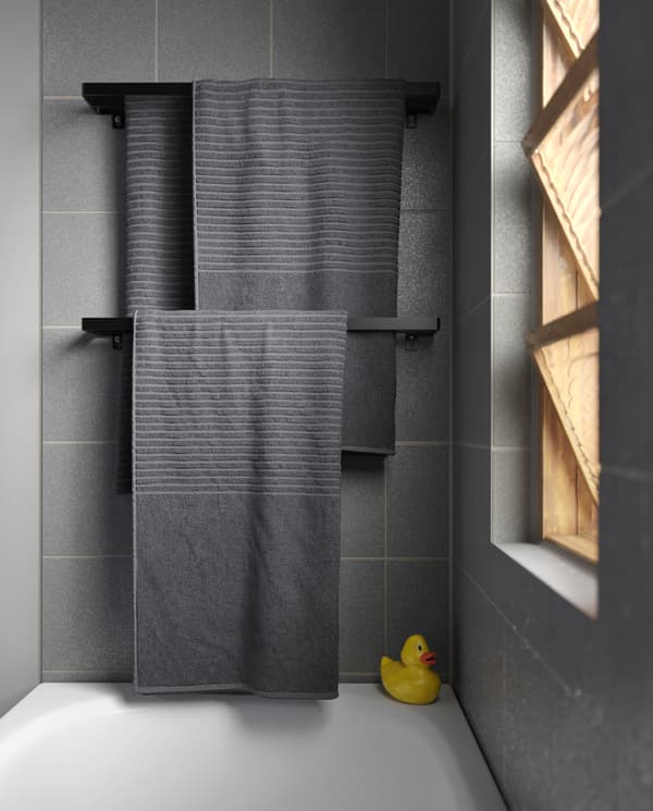 Grey towels hang on two black towels rails that are wall-mounted over a bathtub. A rubber duck stands in the bathtub corner.