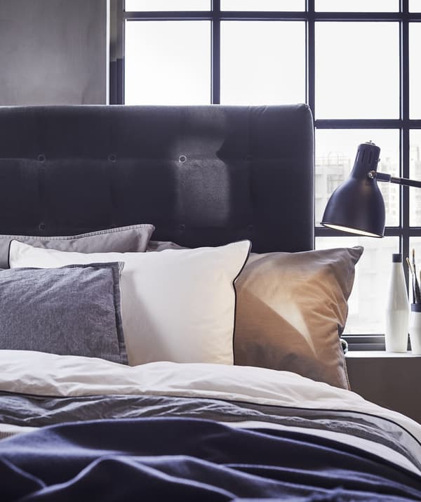 Gros plan d’un lit avec une superposition d’oreillers et de linge de lit en blanc et gris, une tête de lit foncée et une lampe de chevet noire.