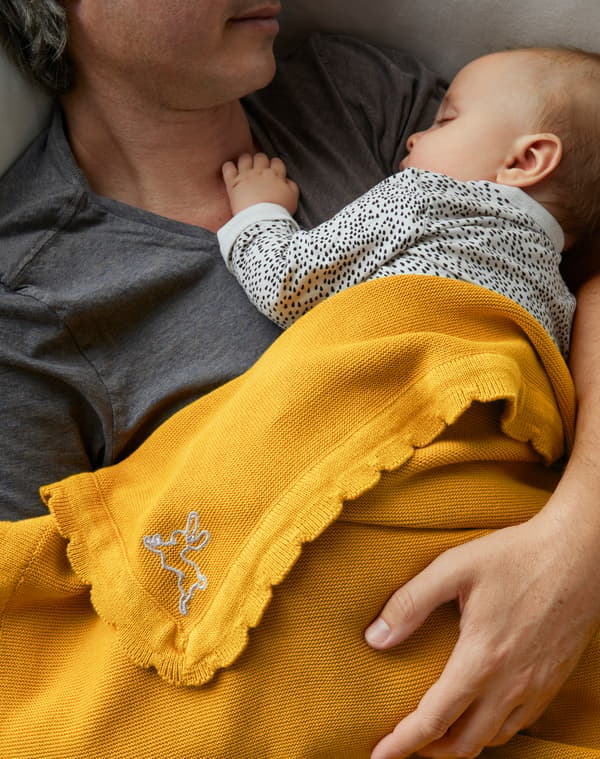 Gros plan d’un père allongé qui soutient d’un bras son bébé endormi sur sa poitrine et de la couverture SOLGUL jaune posée sur eux.