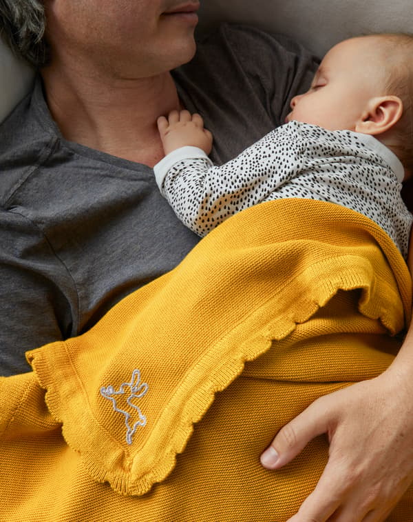 Gros plan d’un père allongé qui soutient d’un bras son bébé endormi sur sa poitrine et de la couverture SOLGUL jaune posée sur eux.