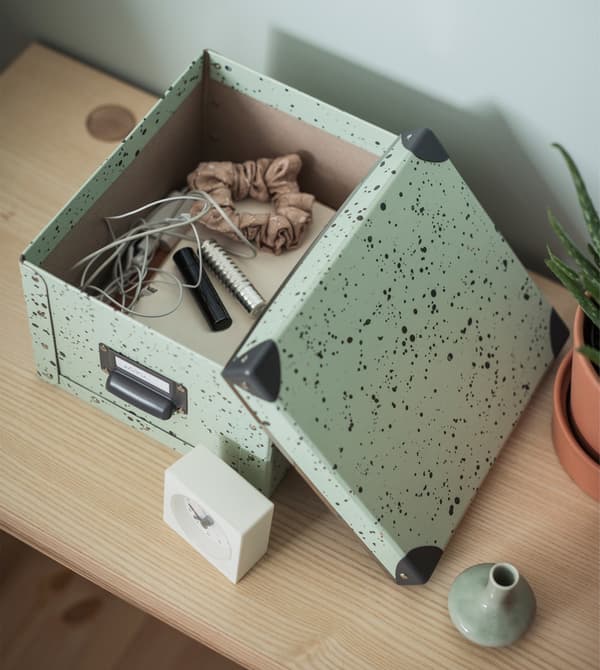 Gros plan d’une boîte de rangement verte FJÄLLA ouverte dans laquelle sont rangés des écouteurs, des bandeaux et d’autres objets.