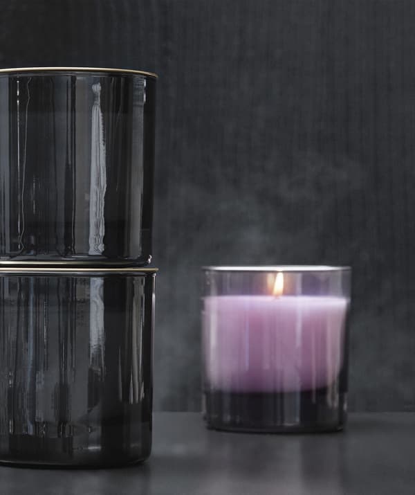Gros plan d'une bougie couleur lavande dans un photophore au verre fumé, sur fond gris foncé.