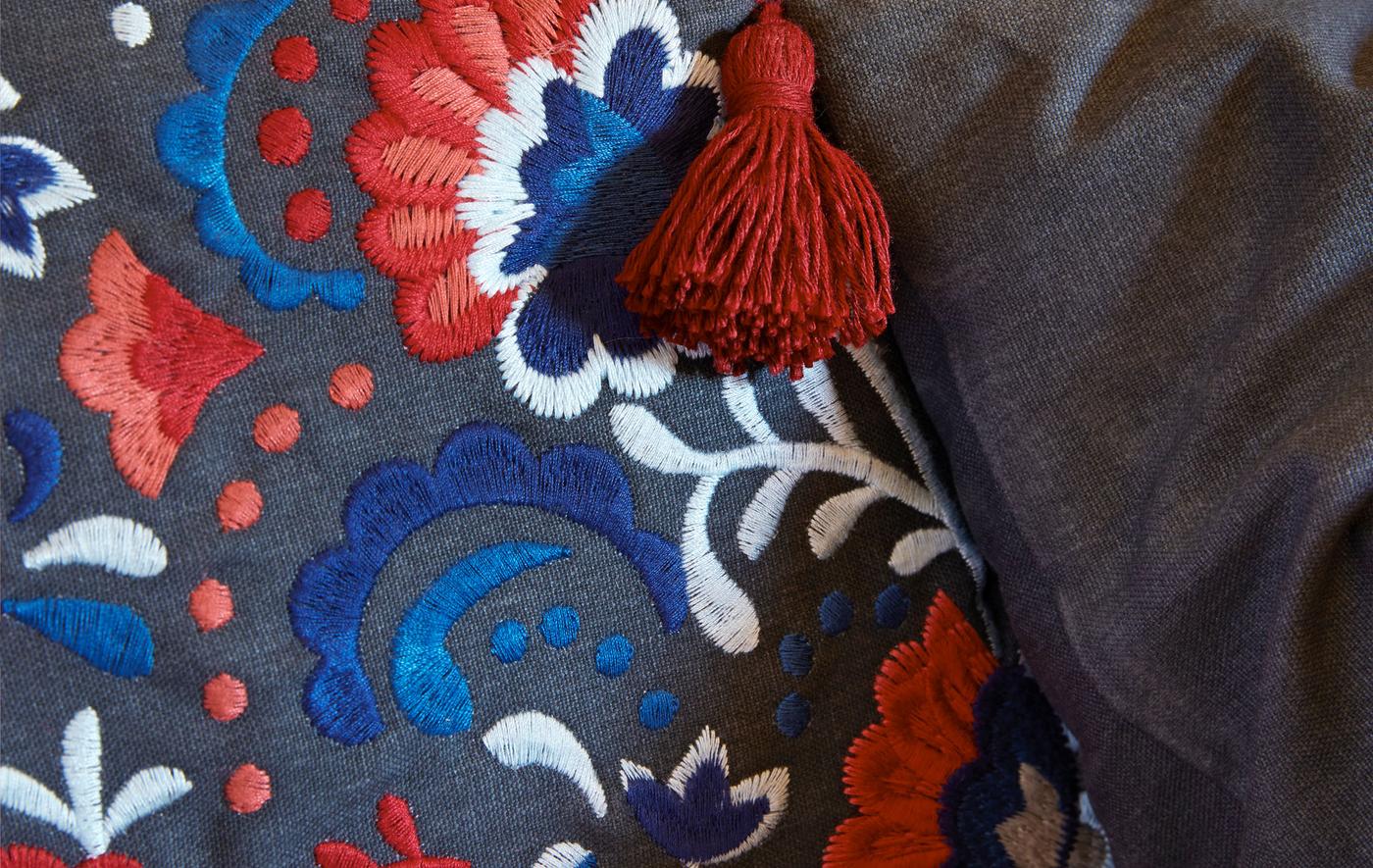 Gros plan d’une housse de coussin, brodée de motifs complexes de différentes couleurs, avec un gland accroché à un coin.