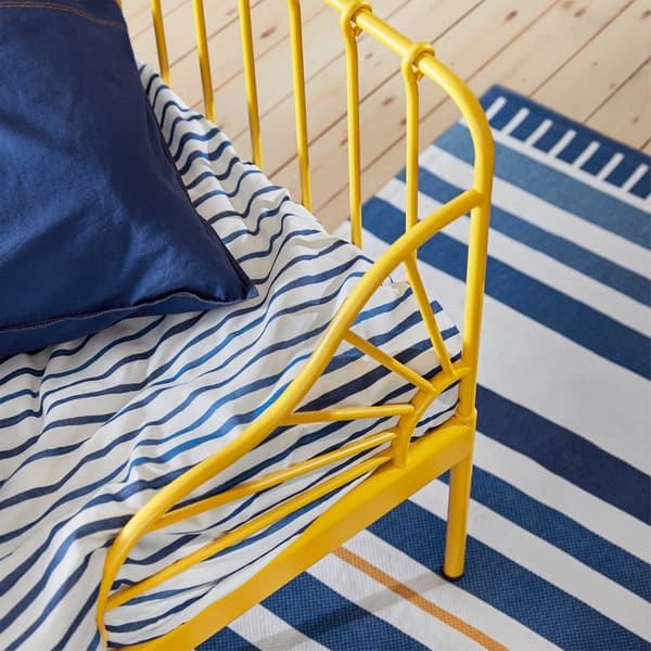 Gros plan sur le coin d’un lit jaune, le linge de lit blanc à rayures bleues et le tapis.