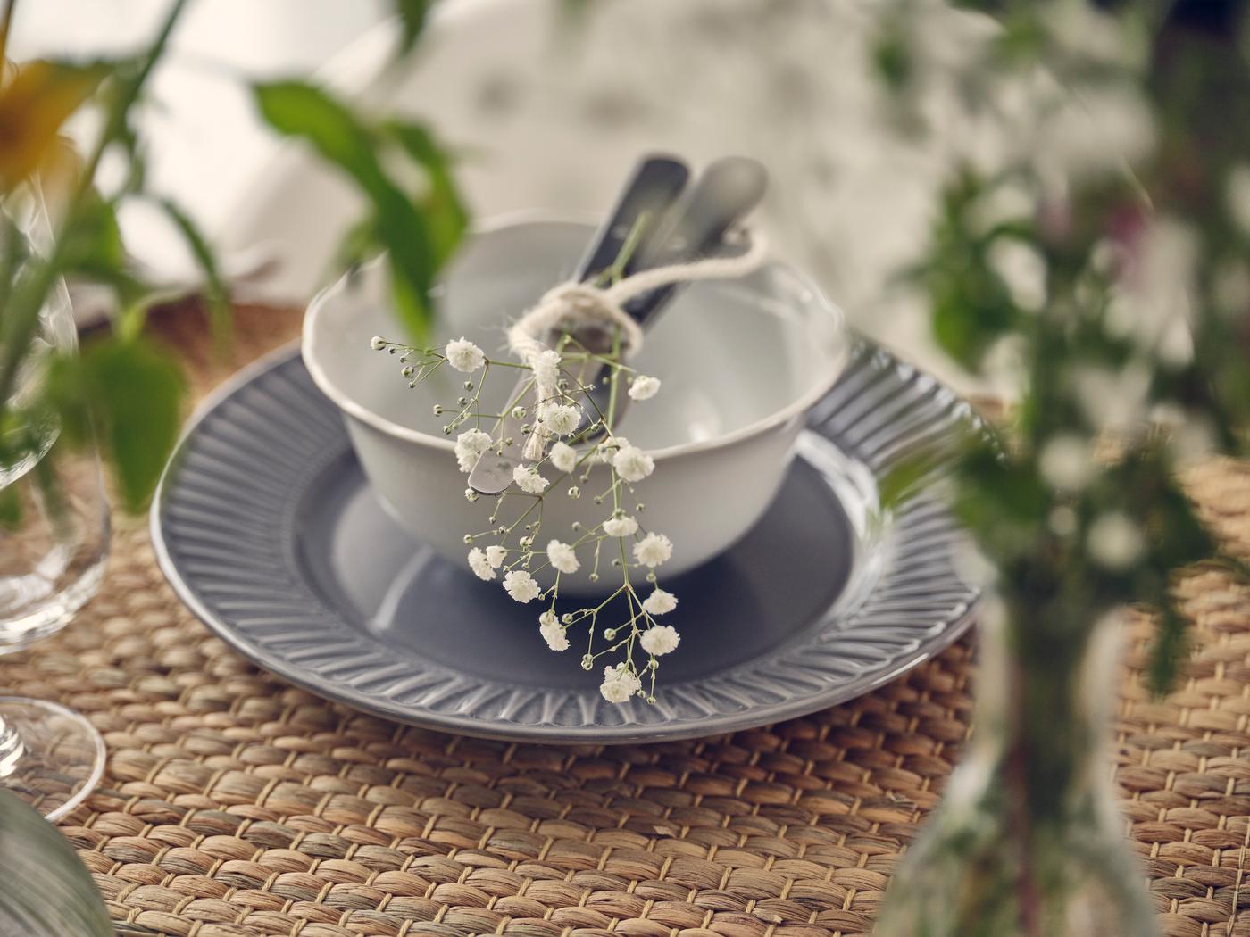 Gros plan sur une table dressée avec un bol blanc, une assiette grise, des couverts et des fleurs délicates.