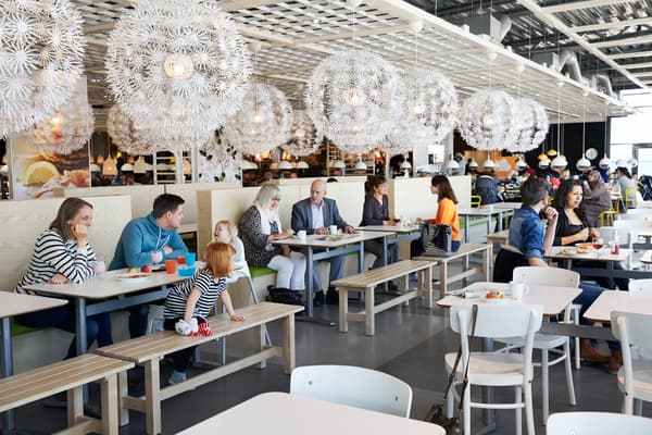 Grupos de familias sentadas en el restaurante de IKEA comiendo. Lamparas blancas en el techo que alumbran y mesas de madera con sillas blancas.