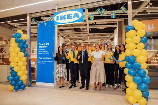 Gruppe von IKEA-Mitarbeitenden steht vor einem neu eröffneten IKEA-Store, umgeben von blau-gelben Luftballons und schwedischen Flaggen. Ein großes blaues Schild mit Informationen hängt links neben dem Eingang.